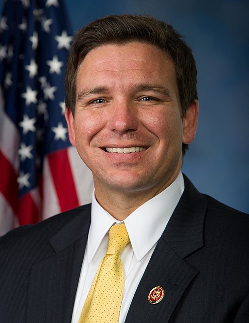 Official portrait of Florida Governor Ron DeSantis in a dark suit and yellow tie, standing before an American flag backdrop.