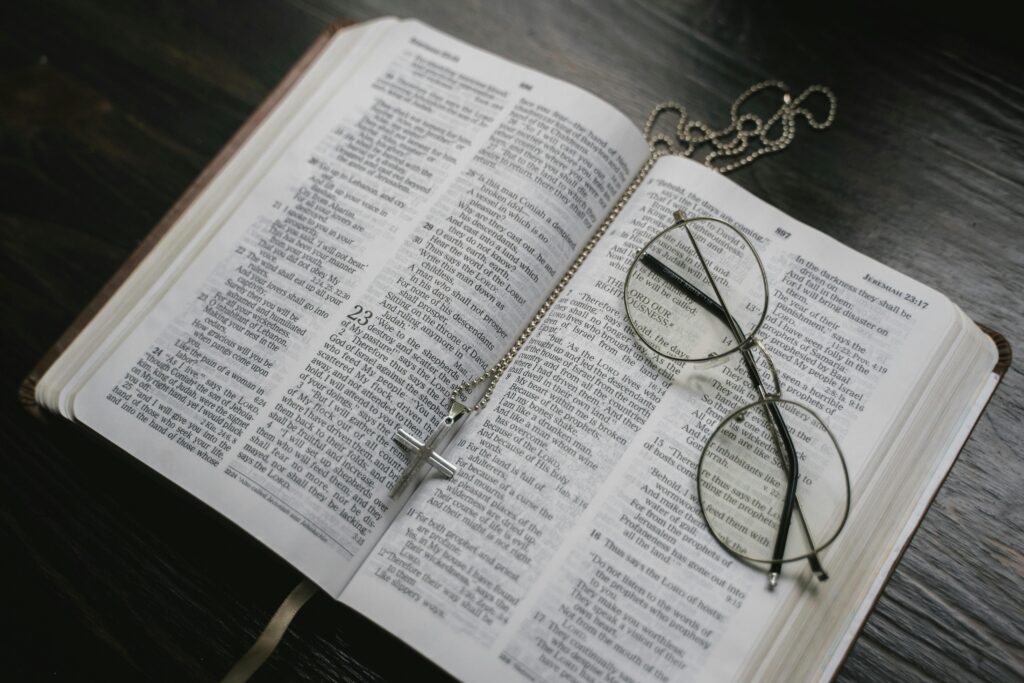 Open Bible with wire-rimmed reading glasses and a silver cross necklace resting on the pages, suggesting scholarly study of religious texts.