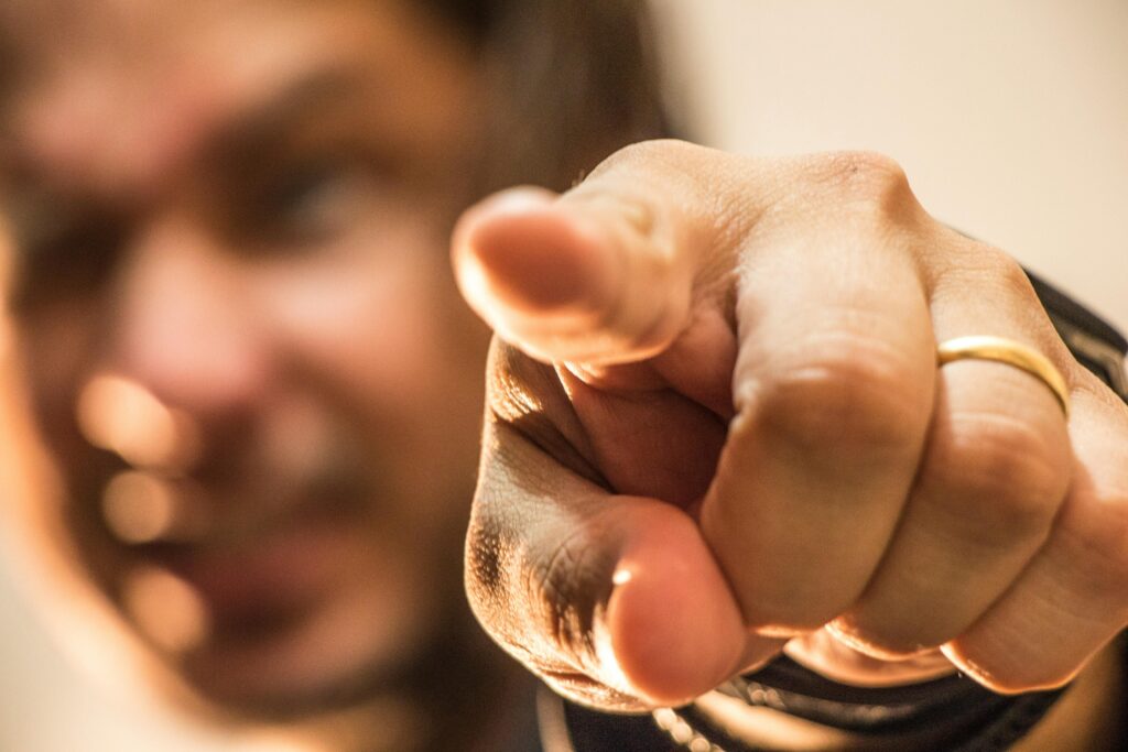 Person points accusingly with finger in foreground, blurred figure behind.