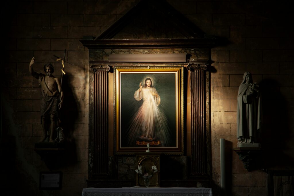 Religious artwork displayed in an ornate church setting, showing Jesus with rays of divine light emanating from his figure, flanked by classical statues.