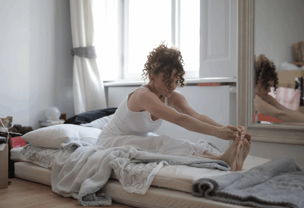 Woman in White Tank Top Sitting on Bed stretching to prevent harm to kidneys