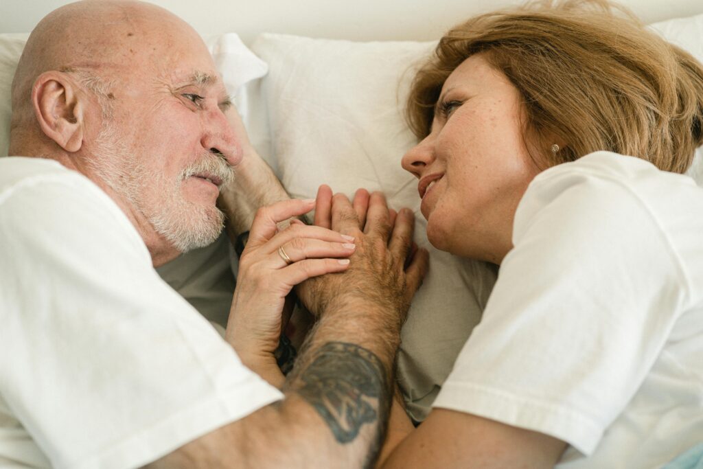 Older couple lying close together in bed, holding hands and gazing at each other with tender expressions