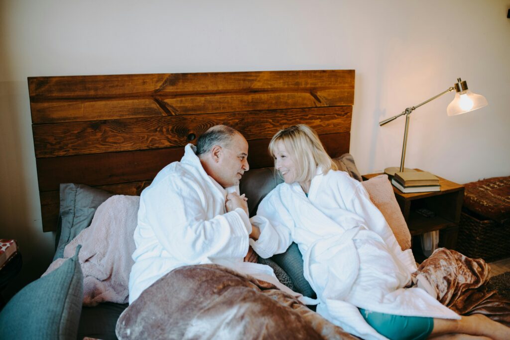 Middle-aged couple in white bathrobes sitting on bed, leaning toward each.