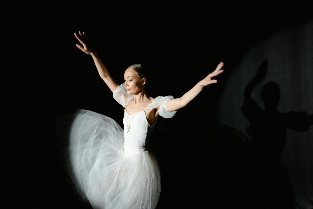 Ballet dancer in white tutu performs with arms raised on dark stage.
