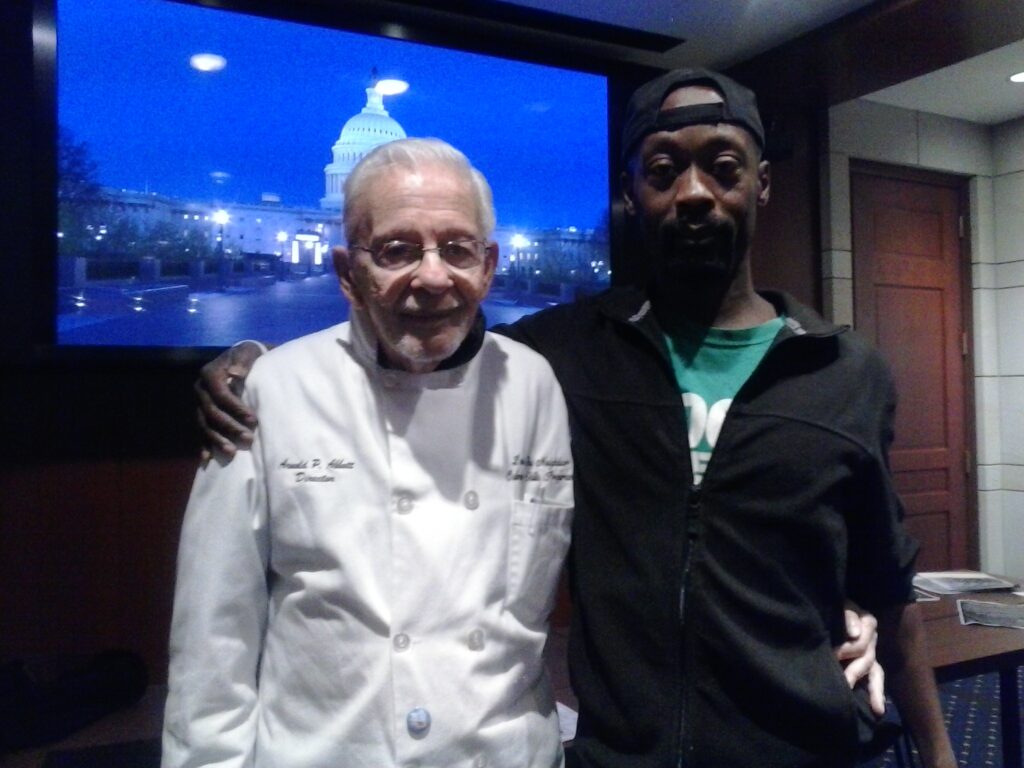 An elderly man in a white chef’s-style coat stands with his arm around a younger man in a black jacket and backward cap. They pose together indoors, with a large screen behind them showing the U.S. Capitol building lit up at night.