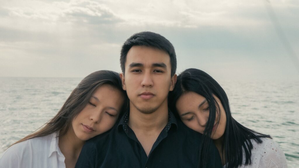 Man stands between two women who lean against his shoulders by water.
