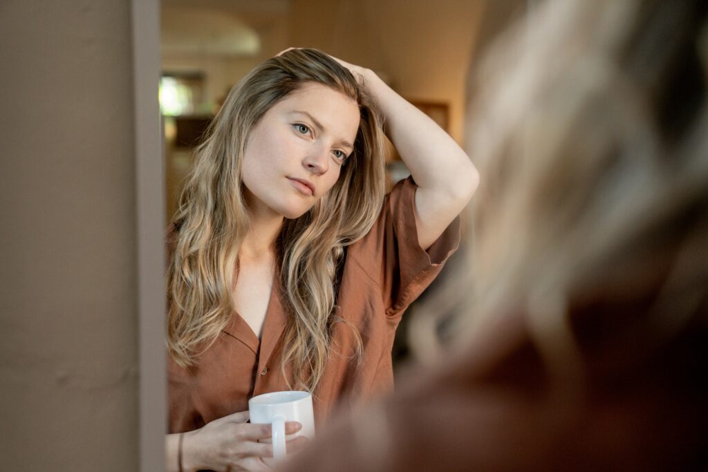 Blonde woman examines herself in handheld mirror with concerned expression.