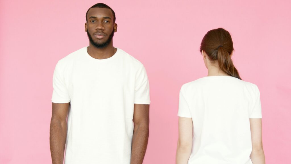 A man facing forwards and a woman facing backwards in white shirts stand next to each other against pink background