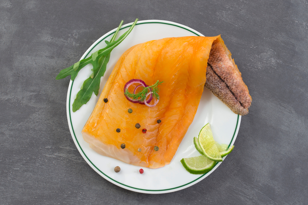 beautiful yellow smoked haddock fish fillet on a plate with a branch of fresh arugula, lime and pink berry on slate background
