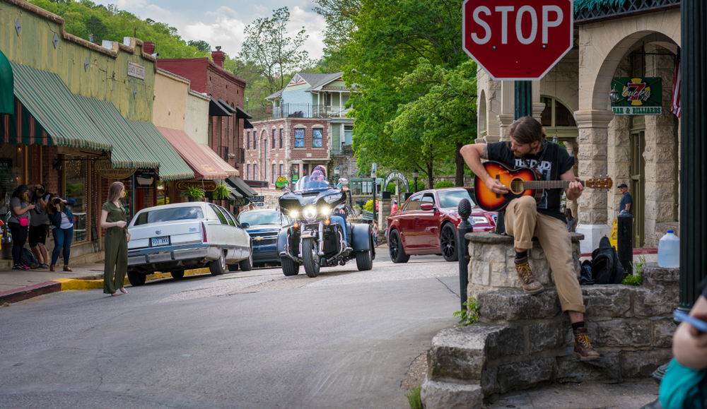 Eureka Springs, Arkansas / USA - April 27 2019: Biker visitors riding motorcycle downtown Eureka Springs, Man playing guitar at stop sign, freedom tourism on 2 wheels, vintage small american town