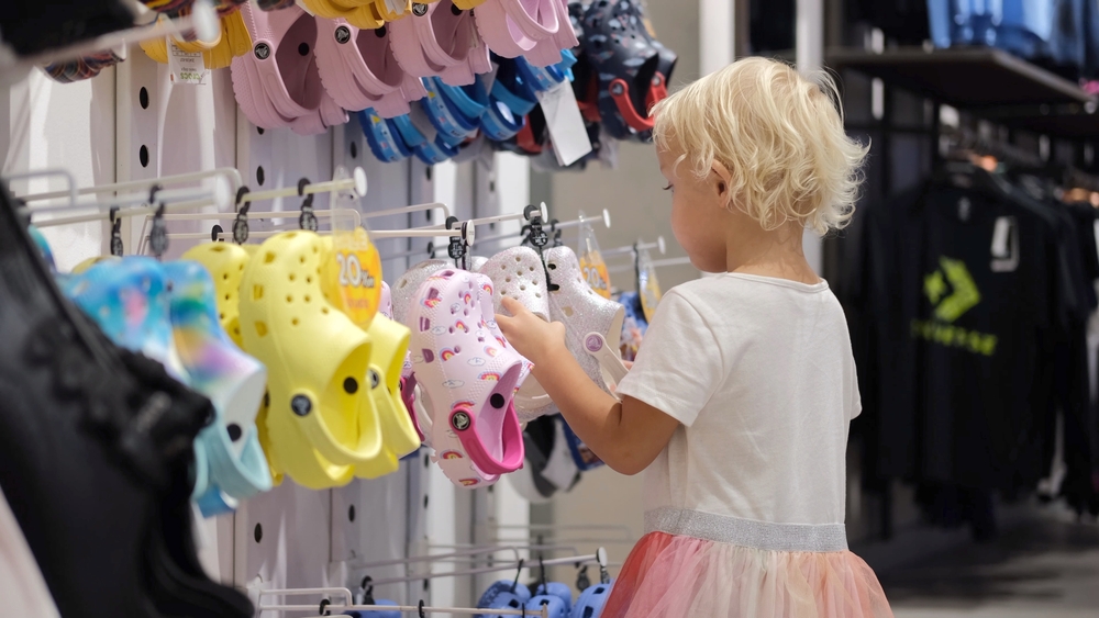 A cute little girl in a shoe store chooses colored slippers for herself. A kid with blond hair shoes that she likes in the market. Children shopping for shoes. Bali-2022-Indonesia