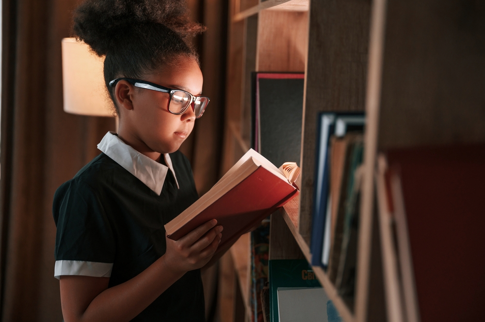 With book in hand. Beautiful light. Cute african american girl in school uniform is at home library.