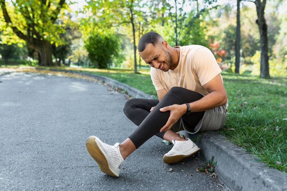 Runner injured his leg while jogging in the park near the trees, hispanic man has muscle spasm massaging his knee, man is running and exercising fitness on a sunny day.