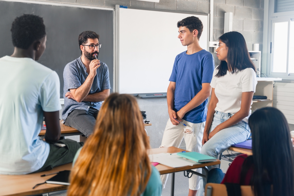 Group of teenagers and male teacher at classroom talking and discussing together

