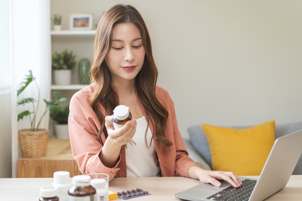 Wellness and dieting asian young woman, girl working from home using computer, typing or searching prescription on medicine label about vitamins information online, holding bottle of food supplement.