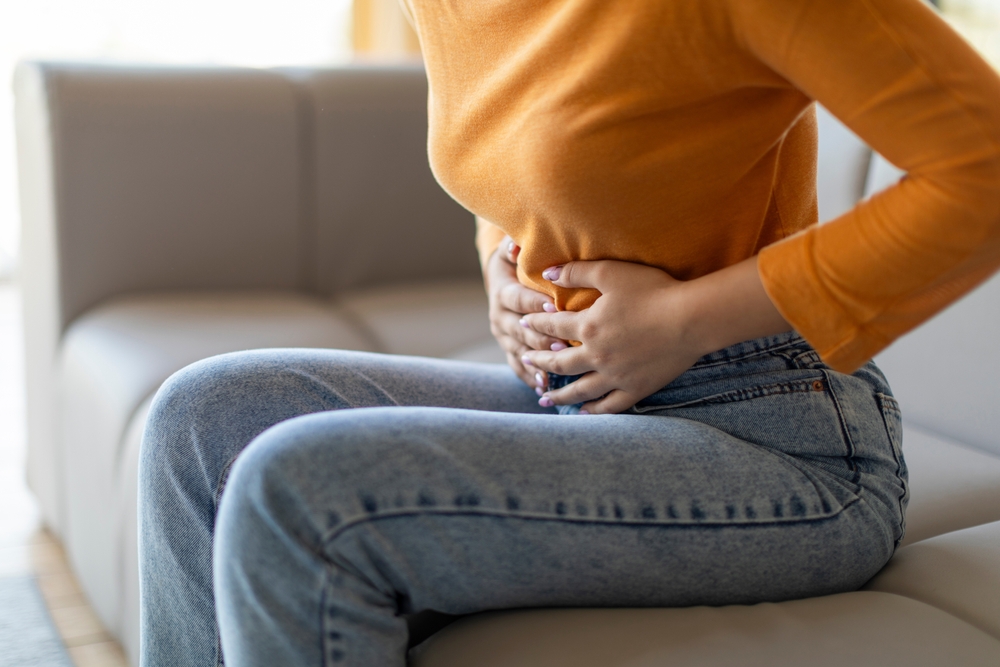 Unrecognizable woman experiencing abdominal pain at home, young female touching her stomach while sitting on couch in living room, showing discomfort and concern, cropped shot, closeup