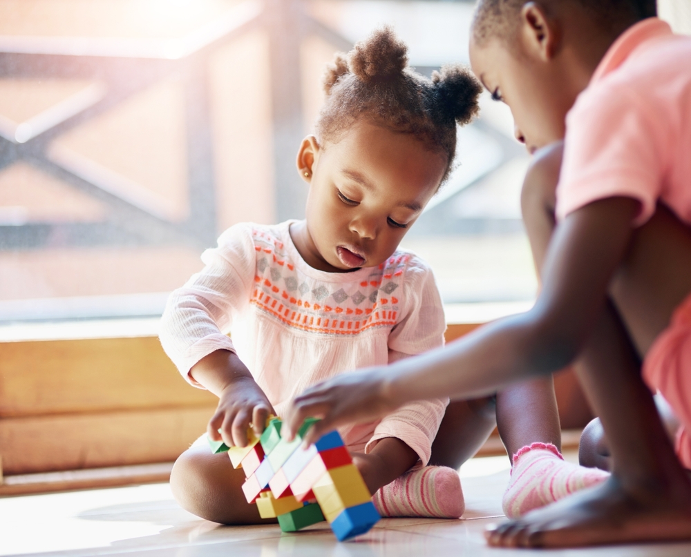 Black child, little girl and siblings playing with blocks or toys together on floor in living room at home. Young African, baby or toddler with playful sister for bonding, help or building at house