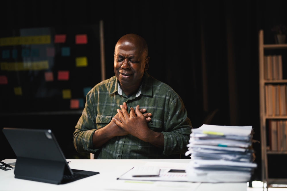 African American man holding chest in pain while working at desk with laptop and documents, highlighting health issues in a professional setting.