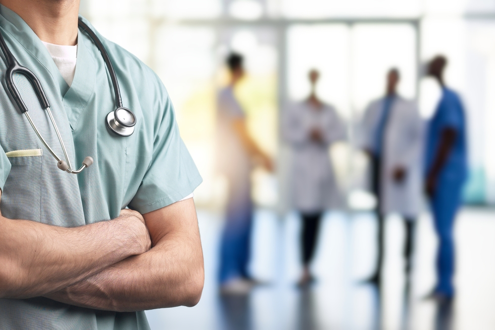 A close-up of a healthcare professional in scrubs with arms crossed and a stethoscope around their neck, with a blurred background of medical staff in a hospital or clinic setting.