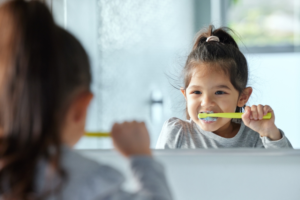 Girl child, mirror and brushing teeth in bathroom for hygiene with wellness, growth and gingivitis prevention in home. Young kid, reflection and tooth cleaning with development, foam and healthy gums