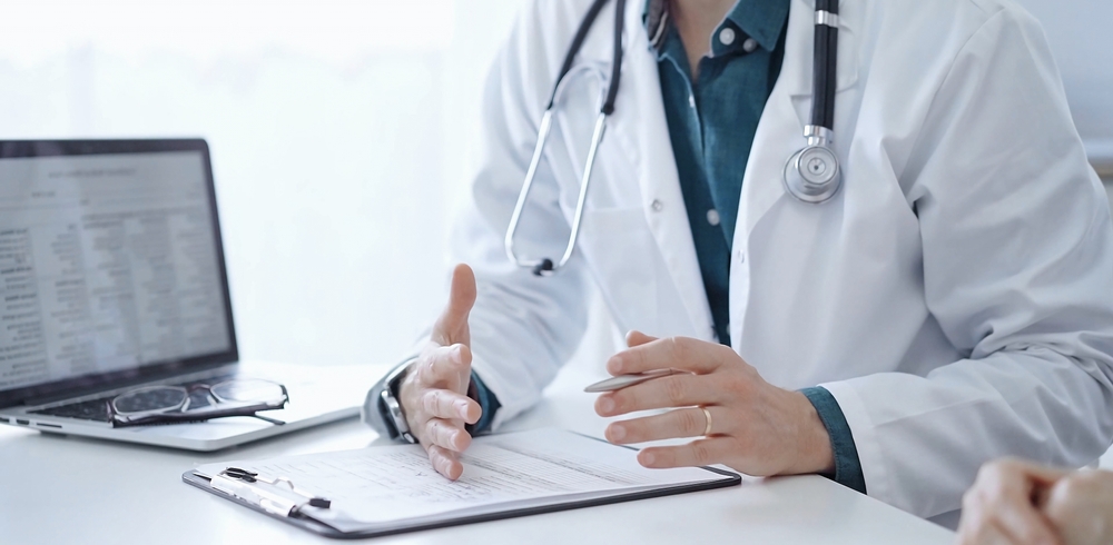 Doctor and a patient. The physician, wearing a white medical coat over a green shirt, is gesturing with his hands during a consultation in the clinic. Medicine