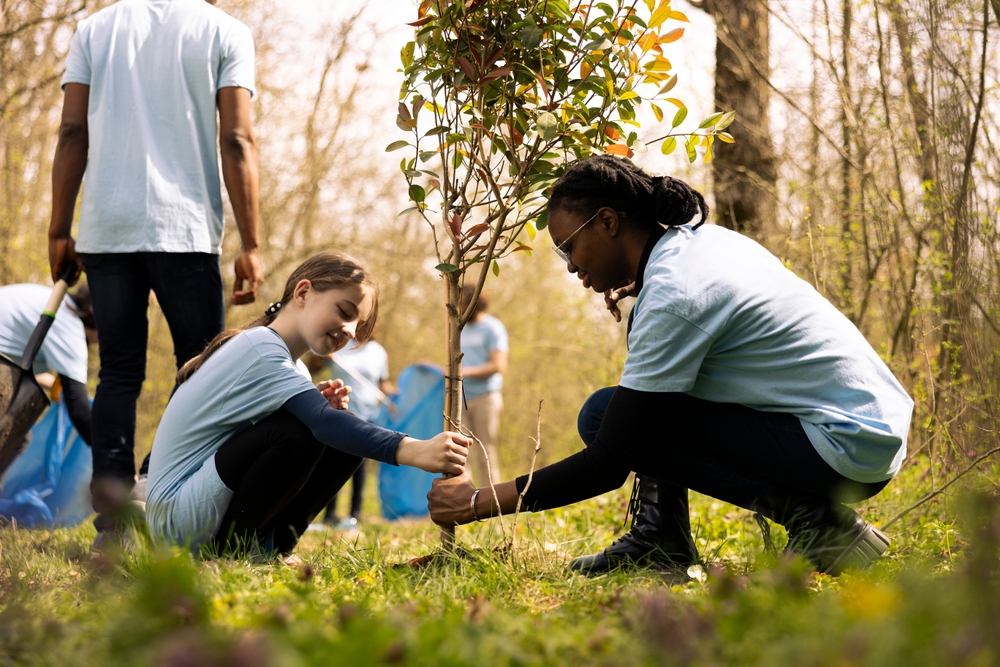 Two diverse activists working together to plant more trees and greenery, filling up ground holes and growing vegetation. Kid and teenager girls collaborating on preserving the environment.