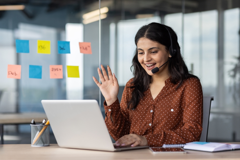 Latin American woman with headset phone using laptop for video call, female online customer support worker waving hand greeting customer, online meeting with colleagues remotely.