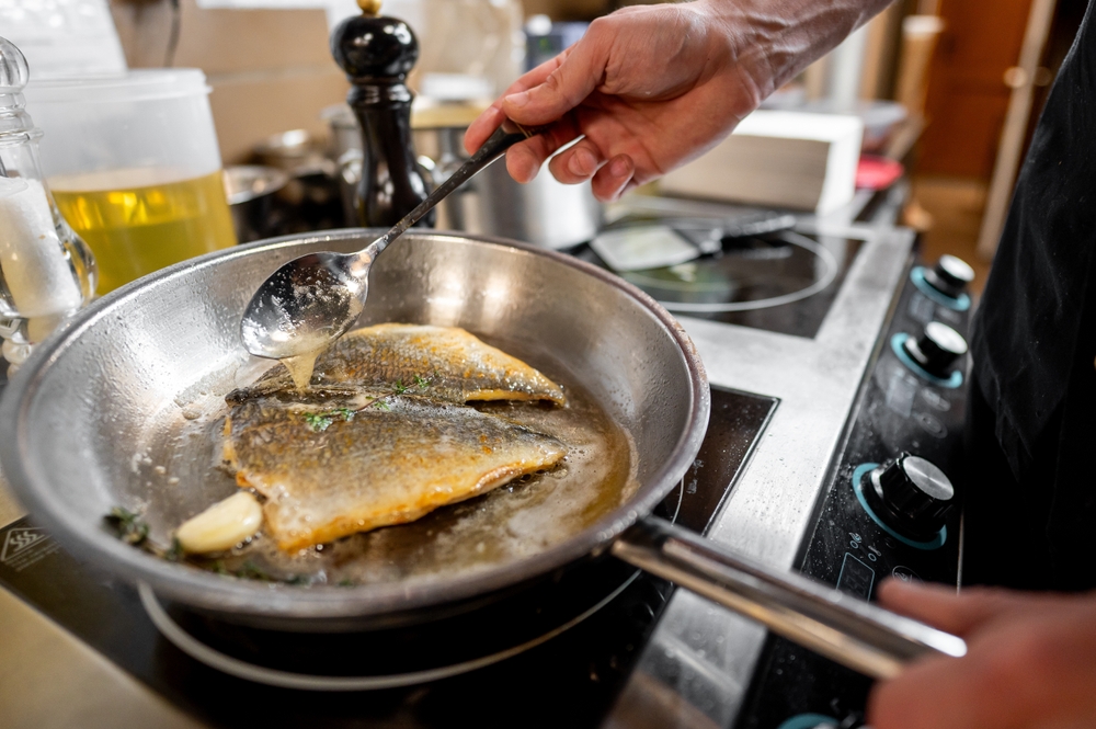 A chef is frying fish in a stainless steel pan, using a spoon to baste it with oil and herbs. The kitchen setting includes olive oil and seasonings, reflecting a culinary vibe.