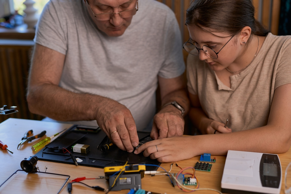 Grandfather and granddaughter work together on an electronics project at a table. Ideal for family, education, and DIY themes