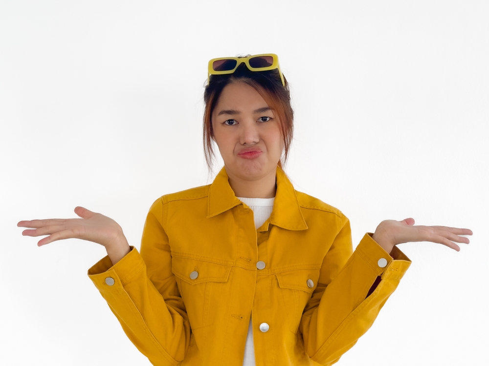 Dislike grumpy face of Asian Thai woman wear yellow jacket and sunglasses, spreading hands none good choice, presenting bad things, standing isolated over white background.