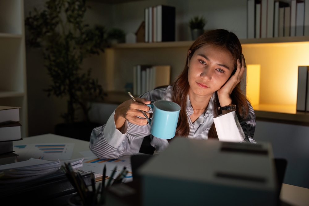 Late Night Coffee Break. Young woman holding a cup while feeling stressed at her desk.