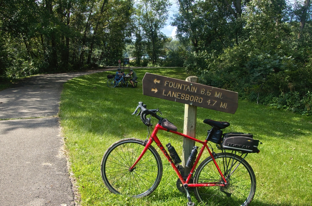 Fillmore County, Minnesota, USA – August 19, 2025: Summer day view of a bike by a mileage sign and cyclists resting along the Root River and Harmony-Preston Valley Trails near Lanesboro, Minnesota.