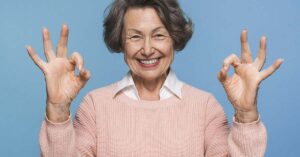 These Personality Traits Are Linked to a Longer Life, Experts Say Elderly woman with her arms raised giving two OK signs with her hands