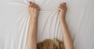 9 Kinds of Orgasms You’ve Probably Never Heard Of (But Might Want to Try) woman reaching up and grabbing the sheets on her bed behind her. Sexual pleasure and orgasm concept