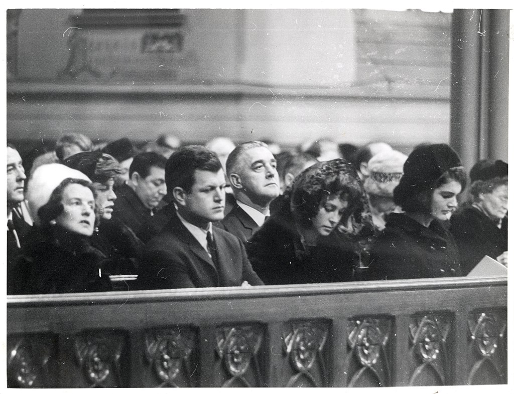 Black and white photo of Rose Kennedy, Senator Edward Kennedy, Joan Kennedy, and Jacqueline Kennedy seated in a church pew wearing black mourning attire with veils.