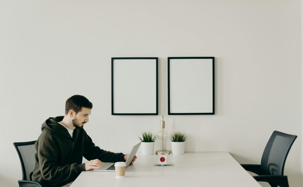 Man in dark clothing writing in notebook at minimalist desk with blank frames on wall.