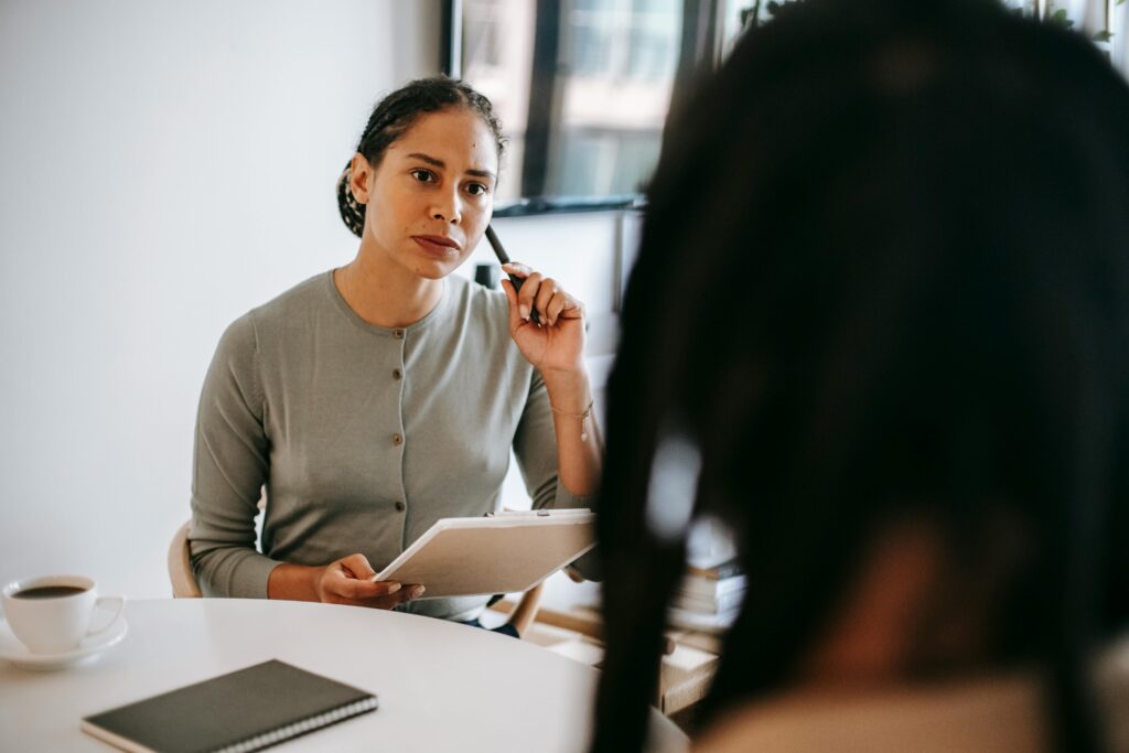 Woman holding pen to cheek with notepad in other hand, looking at another person during conversation.