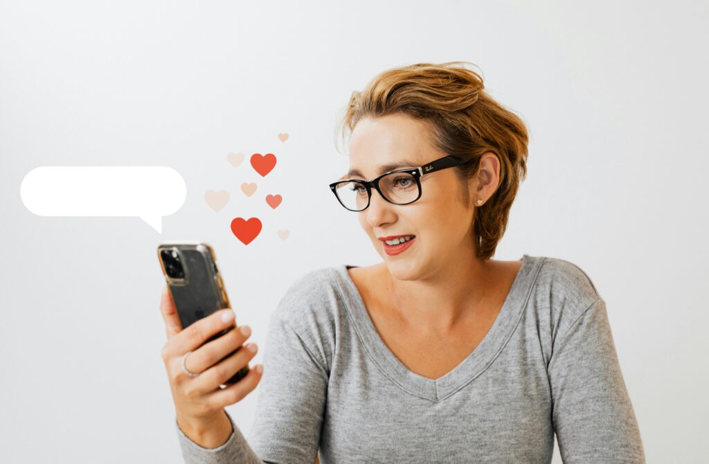 Woman with glasses smiling at smartphone with heart emojis floating nearby.