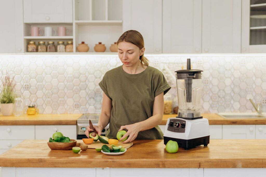 A woman chops avocado and vegetables in a tidy kitchen, with a blender on the table ready for use.