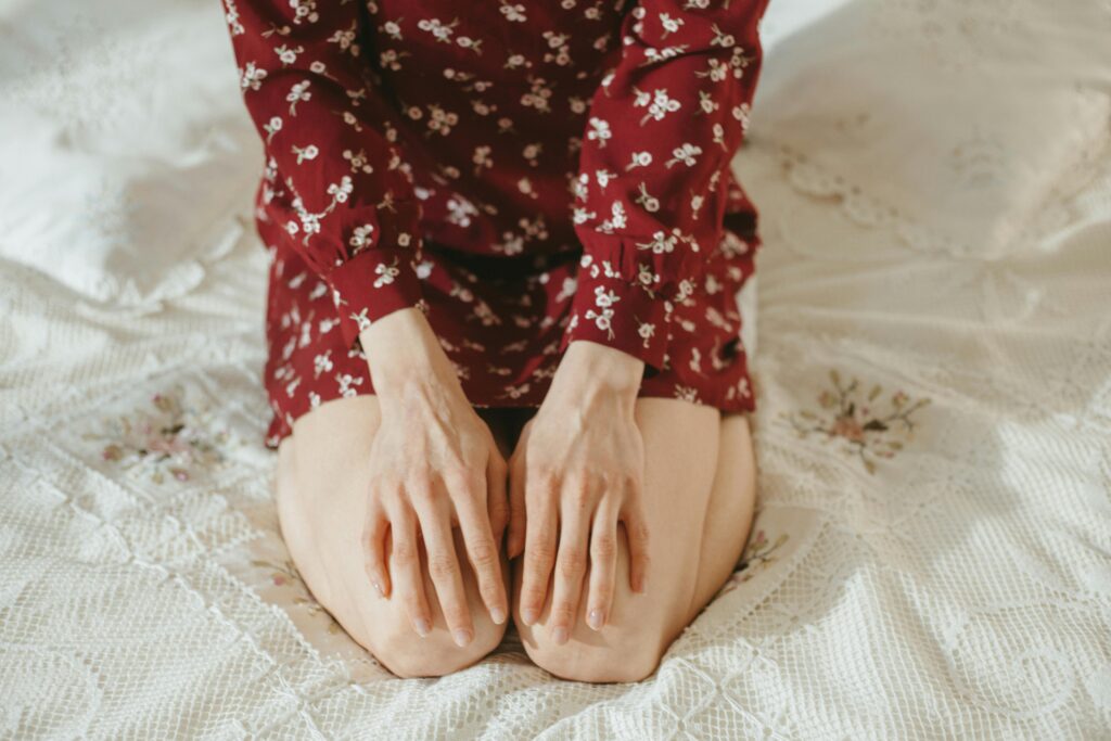 Person in red floral outfit kneels on a bed with hands resting on their thighs.