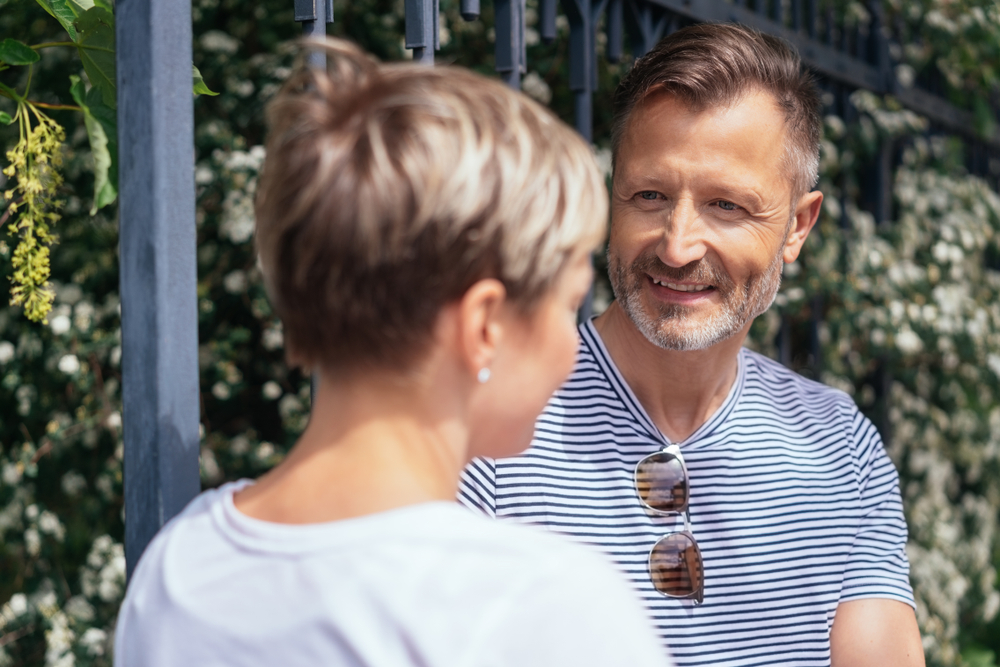 Middle-aged man and woman standing in front of greenery chatting outdoors in the sunshine with over the shoulder view to the smiling man