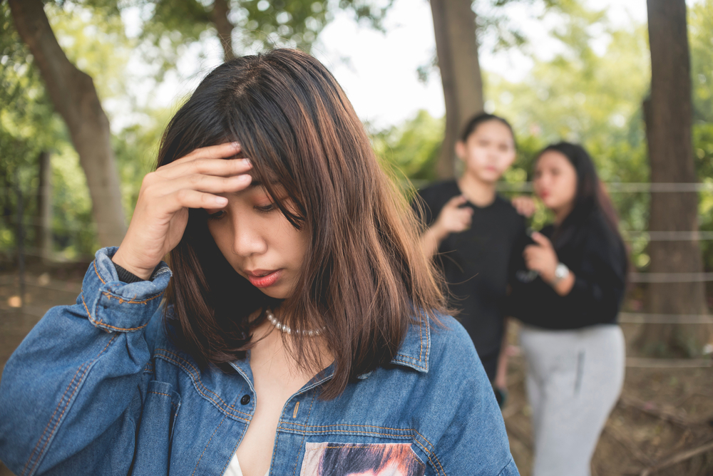 A depressed young woman holds her forehead in shame while 2 women talk and gossip behind her back. Teenage bullying or backstabbing.