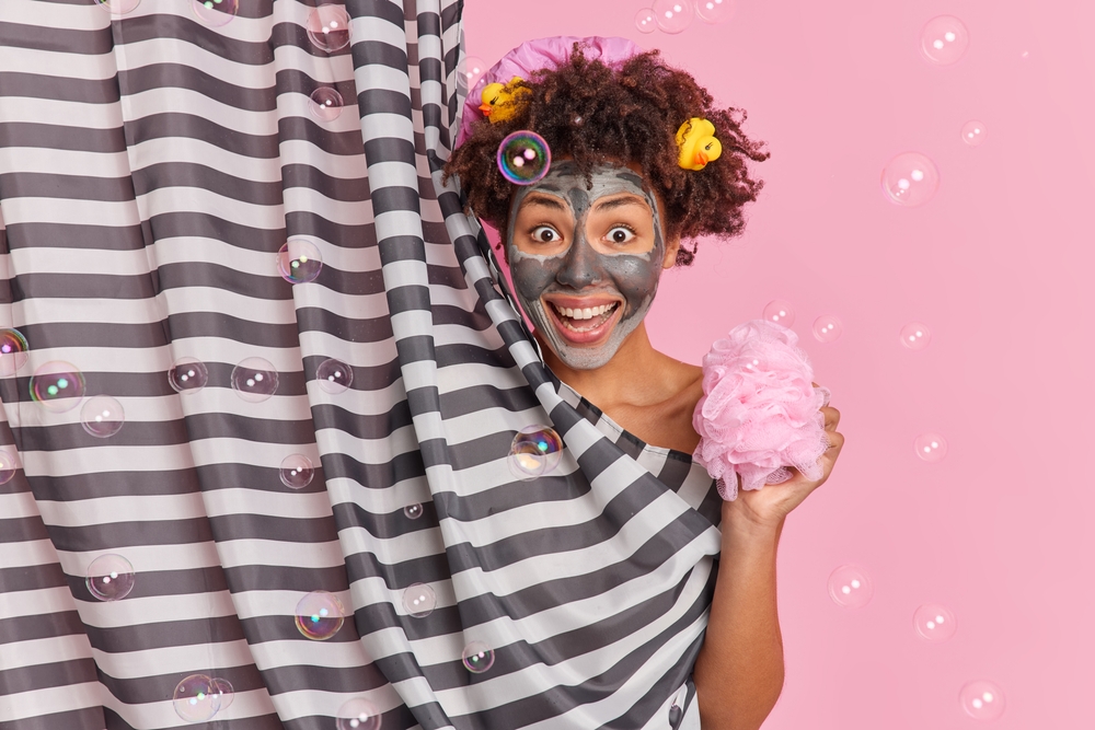Positive curly haired woman with rubber ducks in hair applies beauty wellness mask holds sponge takes shower hides behind striped curtain isolated over pink background with soap bubbles around