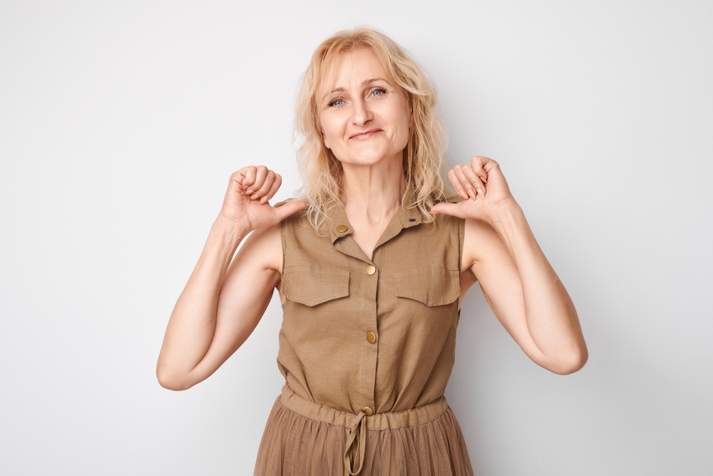 Portrait of blonde mature woman smiling, looking confident, pointing oneself with fingers isolated on white background. Chooses herself, self-esteem concept