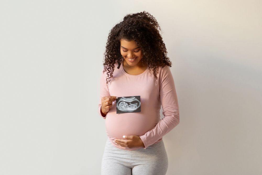 The idea of giving birth. Young Black Woman Holding Ultrasound Image Of Her Baby And Focusing On Her Belly While Standing Against A White Wall At Home, Expectant Mother Keeping A Sonography Scan