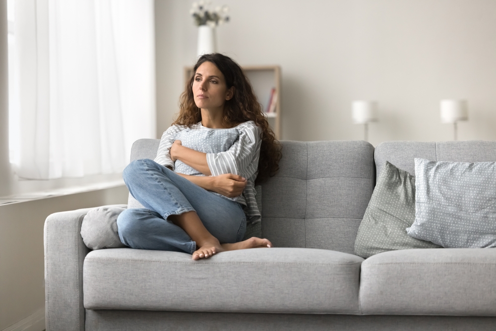 Sad lonely young woman sitting on couch in modern home interior, keeping closed pose, tightening pillow, suffering from depression, apathy after stress, trauma, loss