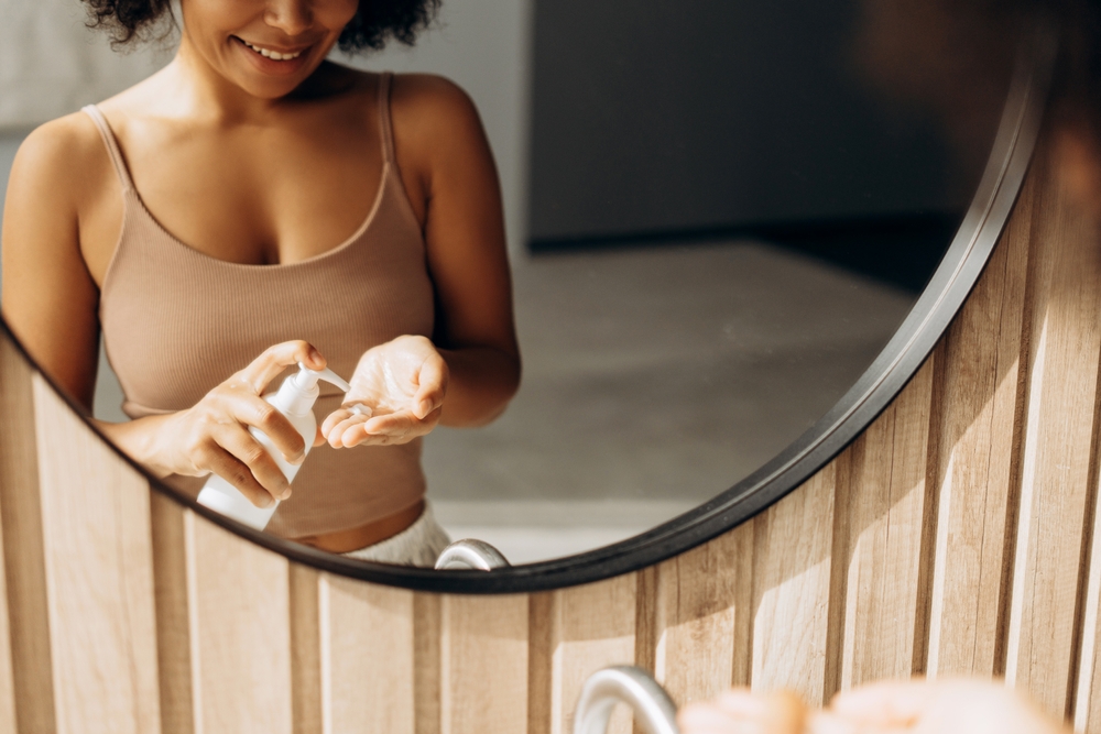 Smiling woman using moisturizing cream or lotion, taking care of her hands skin after shower, standing in front of bathroom mirror