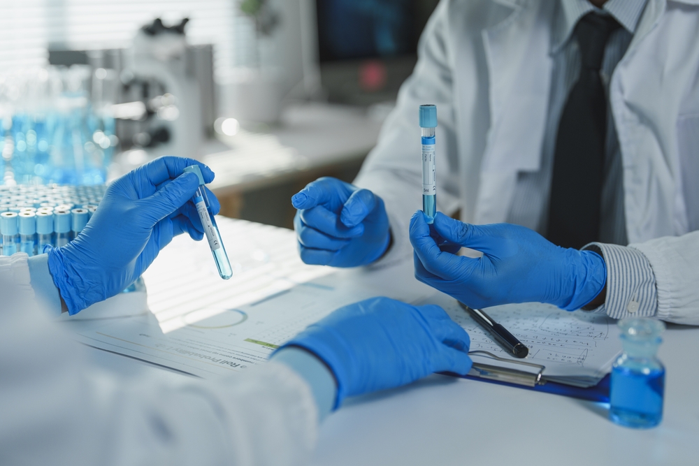 Two scientists wearing blue gloves are comparing test tubes containing blue liquid while reviewing data on a clipboard in a laboratory setting, potentially conducting medical or scientific research