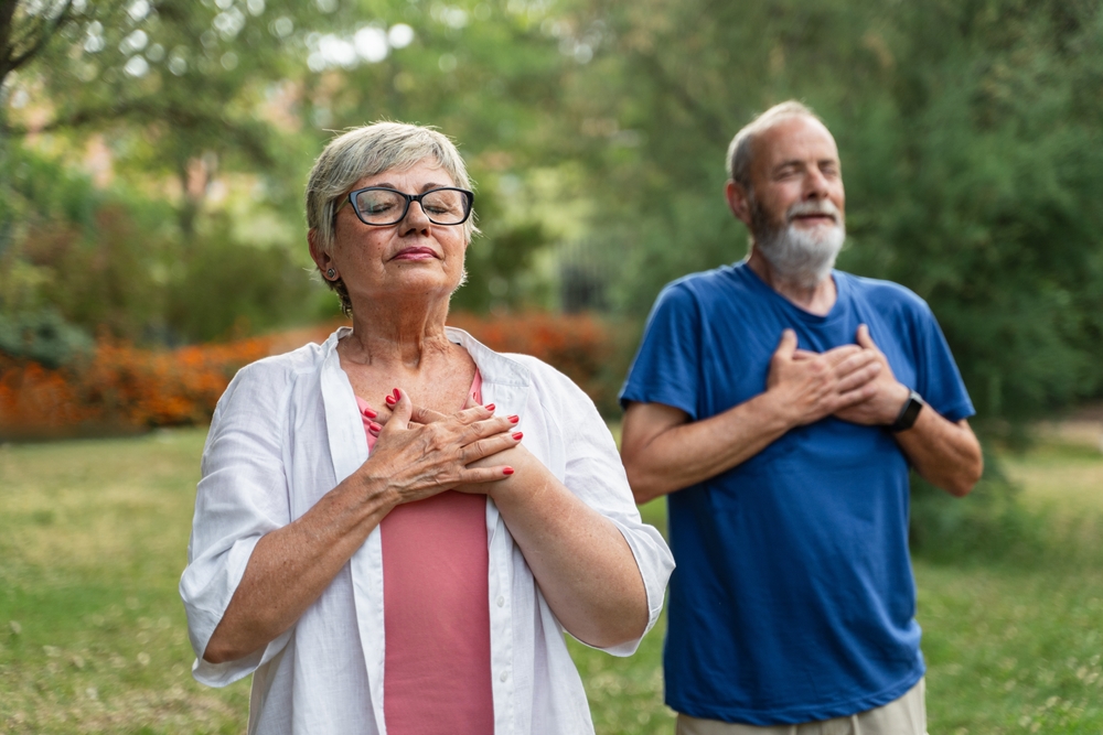 Healthy senior couple holding hands on chest, breathing fresh air, relaxing after yoga stretch, practicing mindfulness and meditation