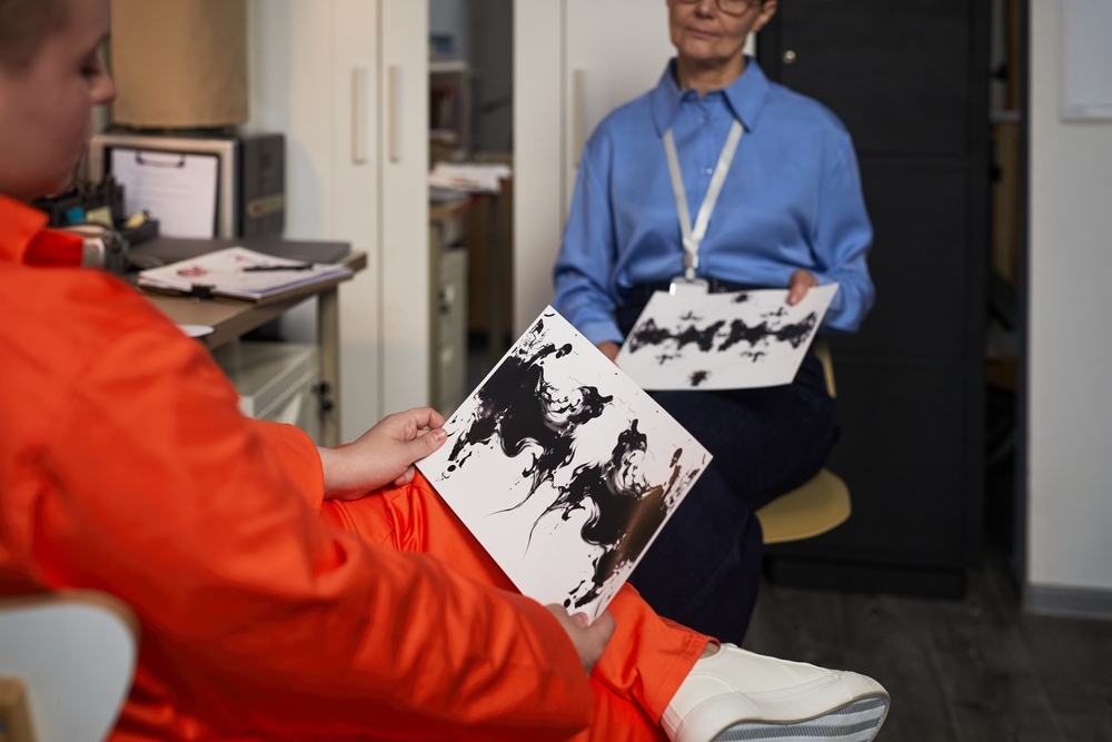 A young Caucasian woman wearing an orange prison uniform sits in an office holding an inkblot test card while an elderly Caucasian doctor examines her during a Prason Therapy Session